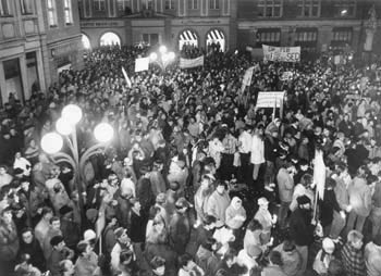 Foto einer Menschenmenge, die sich Abends mit Transparenten auf einem Platz versammelt.