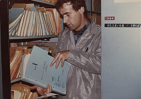 Foto eines Mannes, der eine aufgeschlagene Akte in der Hand hält. Im Hintergrund ist ein Schrank voller Akten zu sehen.