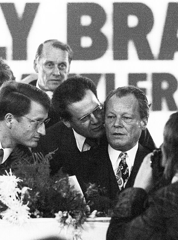 Nahaufnahme Willy Brandts auf einem Podium. Günter Guillaume flüstert dem aufmerksam zuhörenden Brandt etwas in sein rechtes Ohr Ohr.