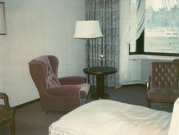 Farbfoto eines Raumes, im Vordergrund ist ein Bett zu sehen, im Hintergrund ein Sessel und ein Stuhl, dazwischen ein Couchtisch, dahinter eine Lampe und ein Fenster.