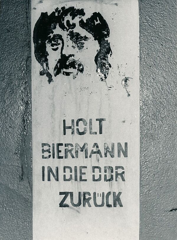 Mit Schablone gesprühtes Graffiti auf einer Wand. Oben ist das Gesicht eines Mannes zu erkennen, darunter der Text "Holt Biermann in die DDR zurück".