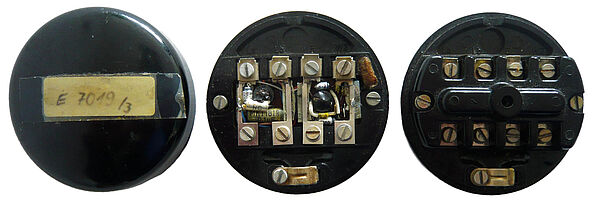 Das Bild zeigt nebeneinander drei schwarze runde Telefonanschlussdosen, die rechte wird von vorne gezeigt, auf ihr klebt ein Aufkleber mit der Aufschrift "E 7019/3". Von der mittleren und rechten sieht man jeweils die Rückseite. In der Mitte der mittleren Dose ist eine kleine elektronische Vorrichtung mit Batterien u. Ä. zu sehen.