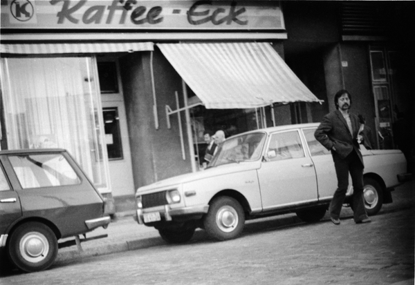 Schwarz-Weiß-Foto eines Mannes der eine Straße überquert. Im Hintergrund sind zwei Autos zu sehen, auf dem Gebäude dahinter steht auf einem Schild über einer Markise "Kaffee-Eck". 