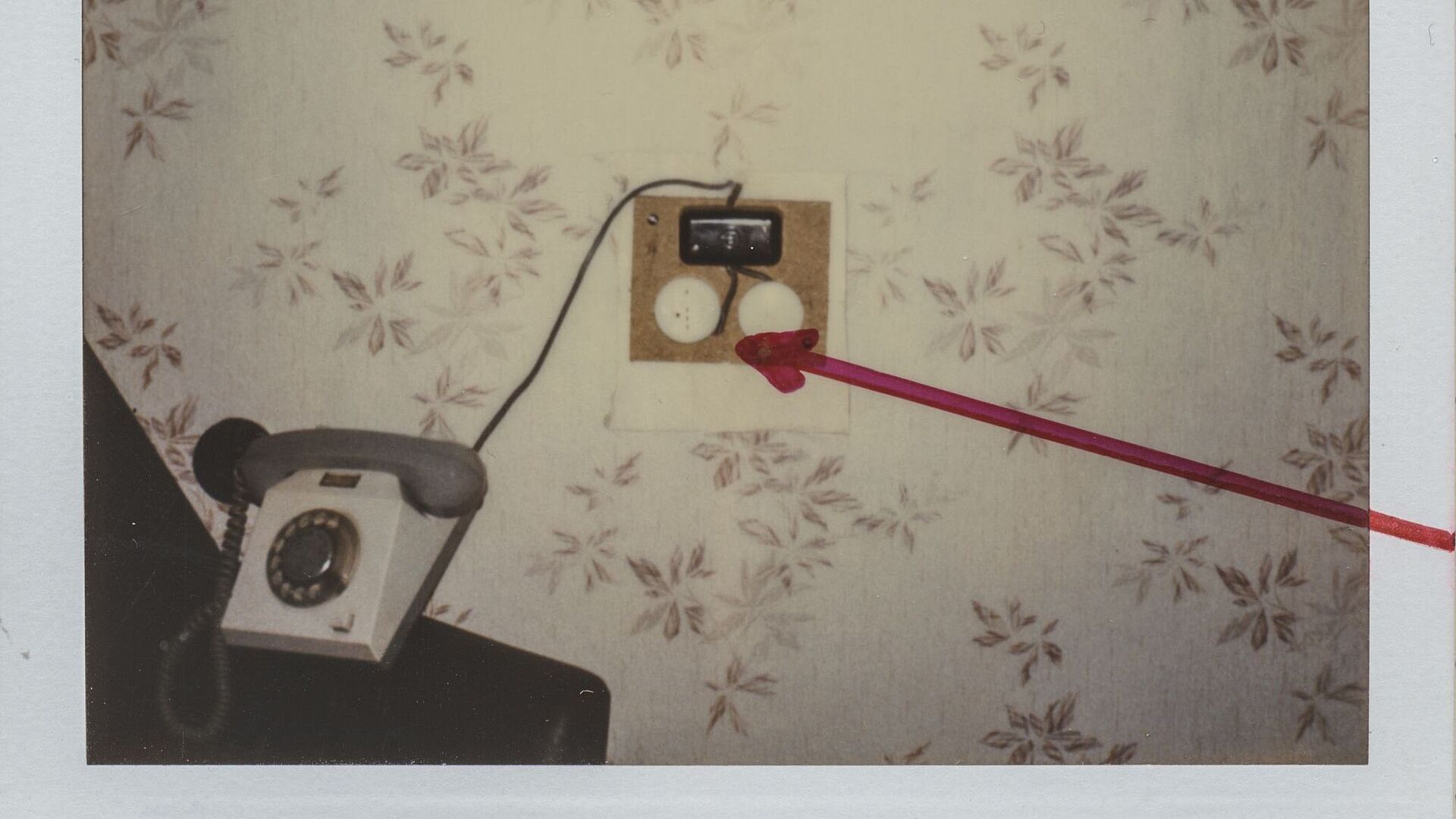Das Polaroidfoto zeigt eine tapezierte Wand vor der ein Telefon steht. Neben dem Telefon ist eine elektronische Apparatur zu sehen. Diese wurde auf dem Polaroidfoto mit einem roten Pfeil markiert.
