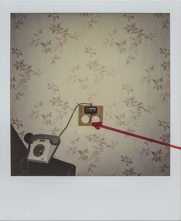 Das Polaroidfoto zeigt eine tapezierte Wand vor der ein Telefon steht. Neben dem Telefon ist eine elektronische Apparatur zu sehen. Diese wurde auf dem Polaroidfoto mit einem roten Pfeil markiert.