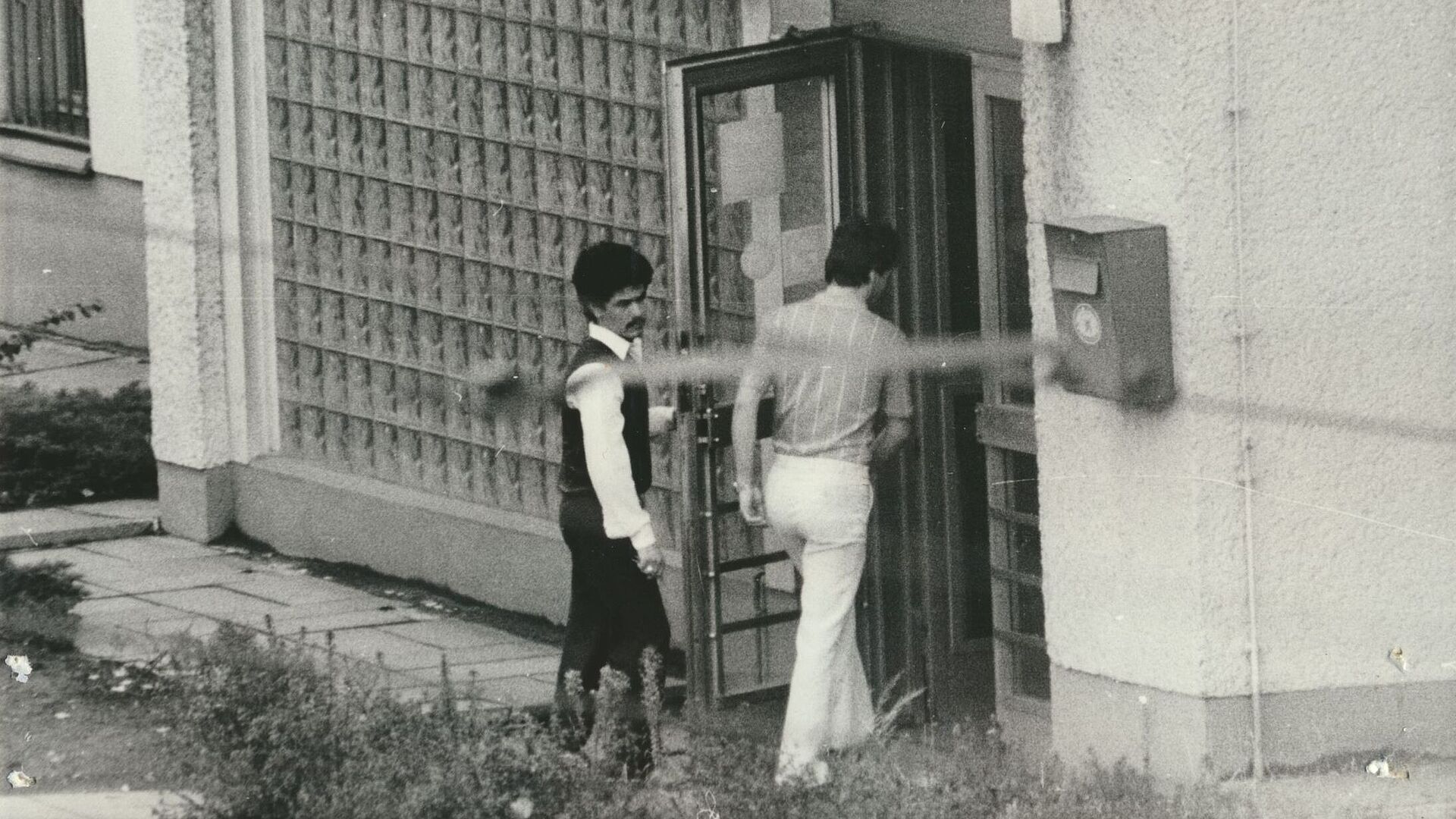 Anscheinend unbemerkt gemachtes Schwarz-Weiß-Foto zweier Männer, die ein Gebäude betreten.