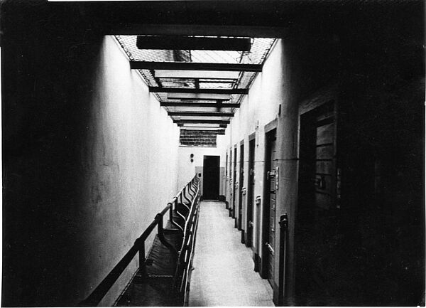 Schwarz-Weiß-Foto eines Gefängnistrakts. AUf der rechten Seite befinden sich Zellentüren, die offene Decke des Ganges ist mit einem Netz verhangen.