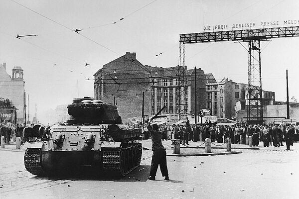 Schwarz-Weiß-Foto eines Mannes, der auf einen Panzer einschlägt. Im Hintergrund ist eine Menschenmenge zu sehen.