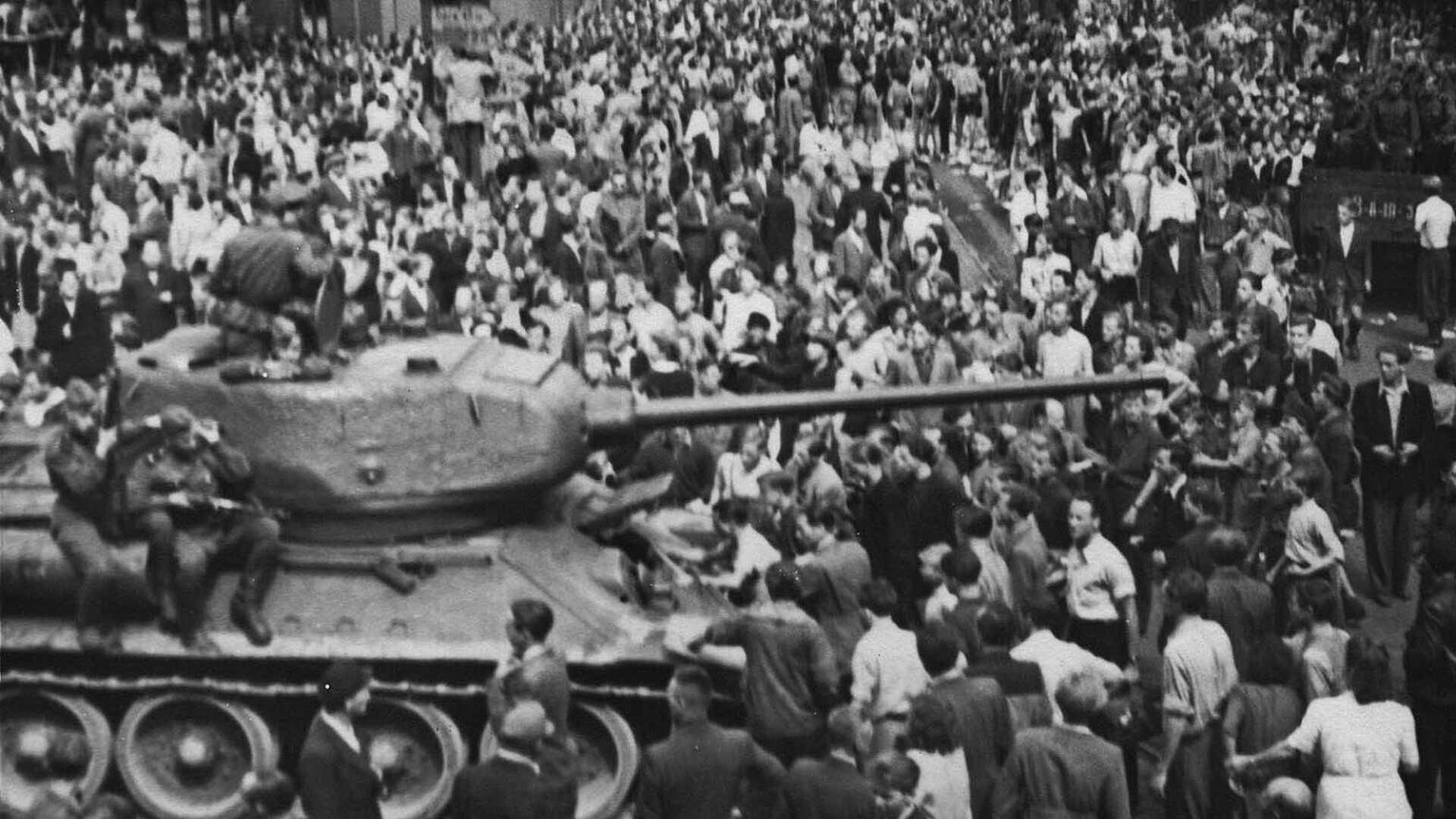 Schwarz-Weiß-Foto einer Menschenmenge, die einen Panzer umringt, auf dem bewaffnete Soldaten sitzen.