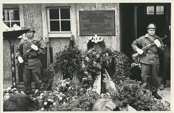 Zwei uniformierte und bewaffnete Soldaten stehen in einem Hof neben einer Plakette, die an die dort stattgefundene Ermordung Ernst Thälmanns erinnert. Vor und neben ihnen ist eine Vielzahl von Blumengestecken und Blumenkränzen.