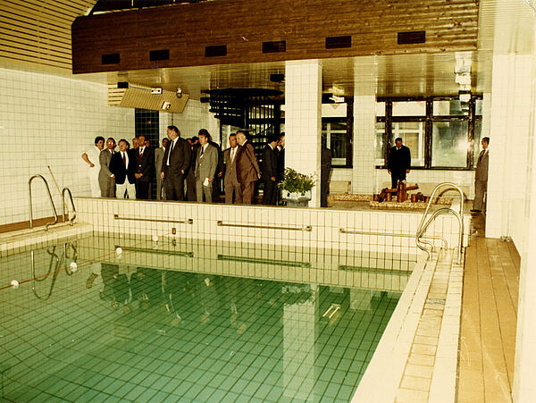 Blick in eine kleine Schwimmhalle in einem neuen Stasi-Hotel über den Pool hinweg auf eine Gruppe von Herren, die sich die Anlage anlässlich der Einweihung des Hotelgebäudes ansehen.