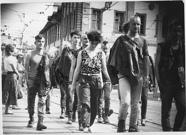 Schwarz-Weiß-Foto mehrerer Personen im Punk-Look auf einer Straße, die an der anscheinend versteckten Kamera vorbei laufen.