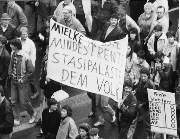 Foto einer Demonstration. In der Bildmitte steht auf einem Transparent "Mielke in die Mindestrente Stasipaläste dem Volk", auf einem rechts daneben ist ein Wahlzettel gemalt, darüber steht "Freie Wahlen".