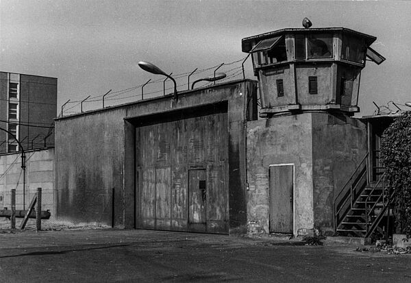 Schwarz-Weiß-Foto des massiven Eingangstores eines Gefängnisses. Rechts daneben ist ein Wachturm, über dem Tor ist Stacheldraht.