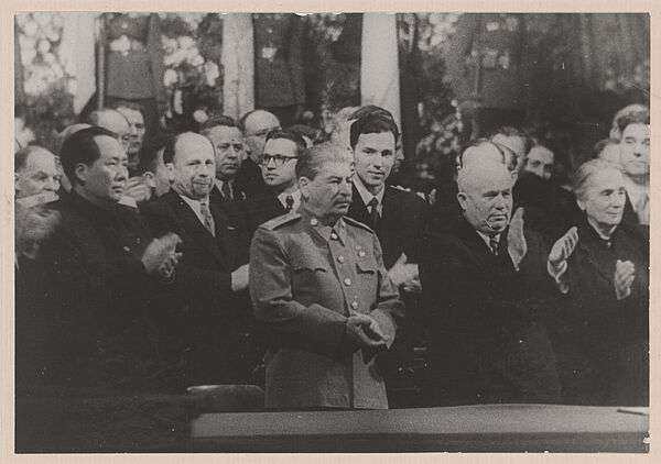 Schwarz-Weiß-Foto mehrerer anzugtragender Personen, die applaudieren. In der Bildmitte ist, in Uniform, Josef Stalin zu sehen.
