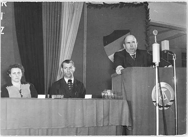 Wollweber steht energisch aussehend auf einer geschmückten Bühne hinter einem Rednerpult. Das Pult ist mit der DDR-Flagge geschmückt. Zu seiner Rechten sitzen ein Mann und eine Frau auf dem Podium 