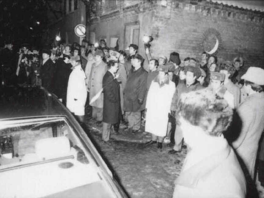 Ankunft der Wagenkolonne mit Bundeskanzler Schmidt und DDR-Staatschef Honecker am Weihnachtsmarkt in Güstrow: Schaulustige winken und halten Blumensträuße hoch, uniformierte Volkspolizisten überwachen die Menge, Pressefotografen machen Fotos. im Vordergrund ist das Heck eines Autos zu sehen