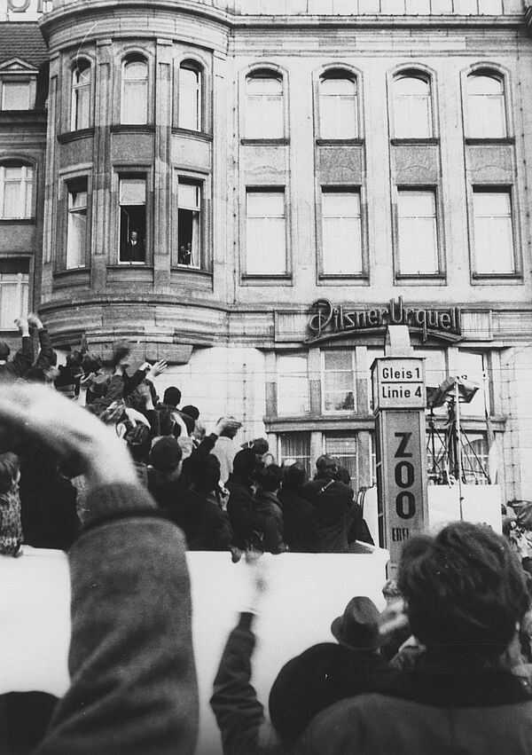 Blick aus einer Menschenmenge heraus auf das Hotel Erfurter Hof. Viele Menschen winken in die Richtung eines geöffneten Fensters im ersten Obergeschoss. Am Fenster steht Willy Brandt.