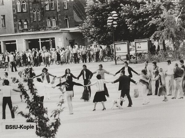 Mehrere Menschen tanzen hädehaltend auf einem Platz im Kreis. Im Hintergrund steht eine Menschenmenge.