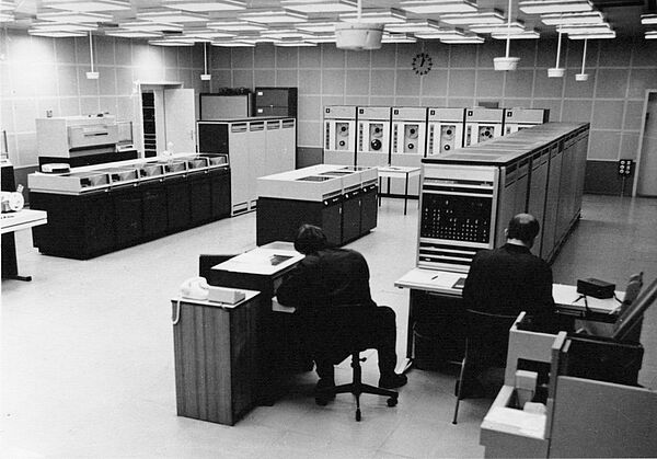 Foto einer Großrechneranlage aus den 1960er-Jahren mit Rechner-, Magnetband und Druckermodulen sowie zwei Operateuren an ihren Arbeitsplätzen.