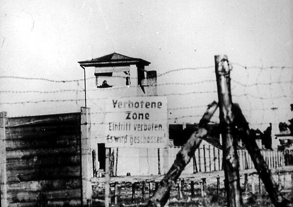 Das Schwarz-Weiß-Foto zeigt im Vordergrund einen Stacheldrahtzaun, dahinter ein Schild auf dem steht: "Verbotene Zone. Eintritt verboten. Es wird geschossen!". Dahinter ist ein Wachturm, auf dem eine Person zu erkennen ist.