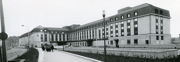 Schwarz-Weiß-Foto eines mehrstöckigen Gebäudes, auf dem Gehweg davor befindet sich eine Personengruppe.