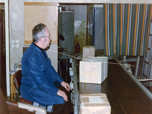 Innenraum: ein von seiner rechten Seite fotografierter etwa fünzig Jahre alter Stasi-Mitarbeiter sitzt mit einem blauen Arbeitskittel bekleidet vor einem kleinen Förderband auf dem Pakete vor ihm entlangbefördert werden.
