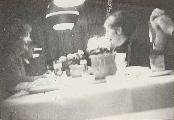 Unscharfes, anscheinend heimlich aufgenommenes Schwarz-Weiß-Foto zweier weiblicher Personen die an einem Tisch sitzen und sich lachend unterhalten. Am rechten Bildrand sind die zwei Hände einer weiteren Person zu erkennen..