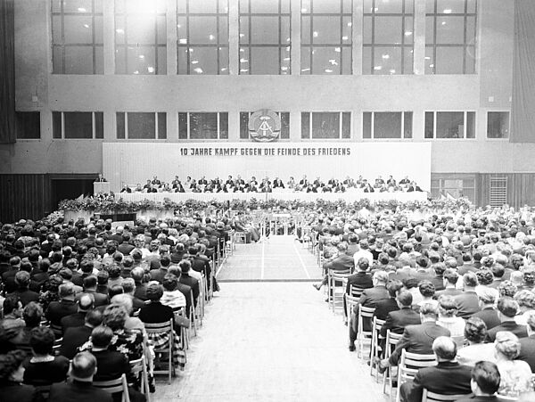 Blick durch eine schlichte, große Halle mit zahlreichen auf Stühlen sitzenden Zuschauern. Ihre Stühle sind auf ein großes, mit zahlreichen Personen besetztes Podium gerichtet. Hinter dem Podium die Losung: „10 Jahre Kampf gegen die Feinde des Friedens“. Darüber das Wappen der DDR.