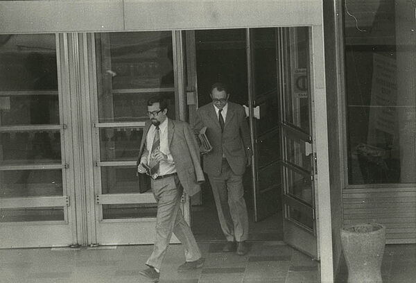 Schwarz-weiß-Foto zweier anzugtragender Männer, die den Eingangsbereich eines Gebäudes verlassen.