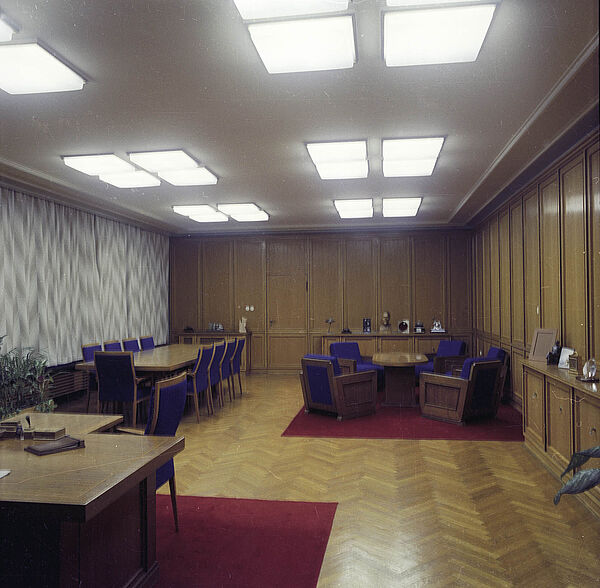 Blick in einen großen Büroraum: vorne links im Vordergrund die rechte Ecke eines Schreibtischs, hinten rechts eine Sitzgruppe mit Tisch und Sesseln, links ein Konferenztisch mit Stühlen. Alle Sitzmöbel haben blaue Bezüge.