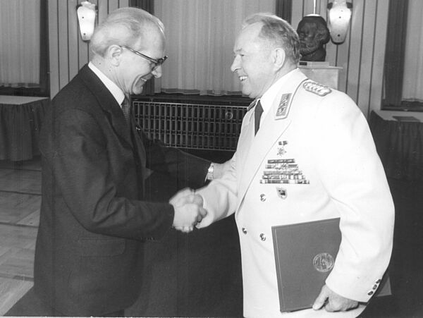 Honecker und Mielke schüttel sich freudig lächelnd die Hände. Honecker trägt einen schwarzen Anzug, Mielke eine weiße Galauniform.