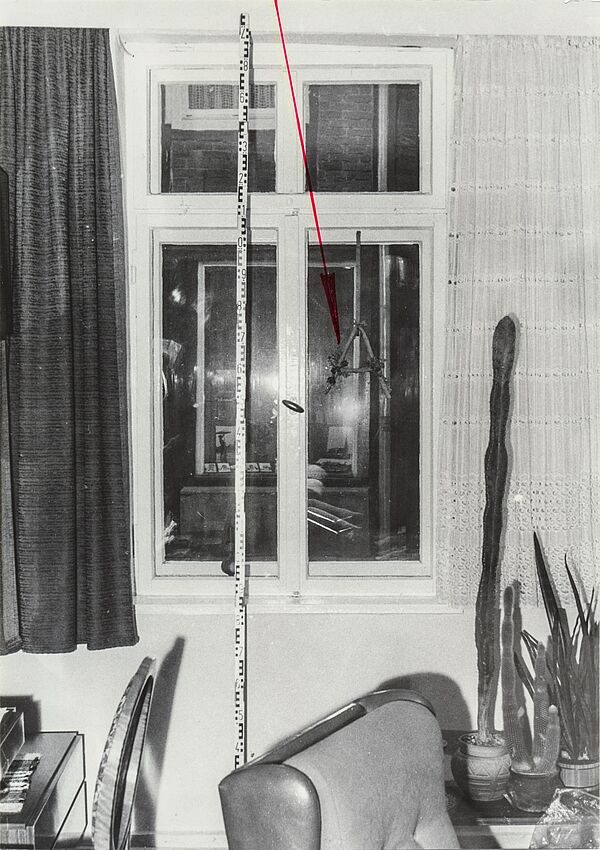 Schwarz-Weiß-Foto eines Fensters in einem Wohnzimmer. Ein eingezeichneter roter Pfeil zeigt auf ein gebasteltes "A", das am Fensterrahmen hängt. Daneben ist ein Maßband.