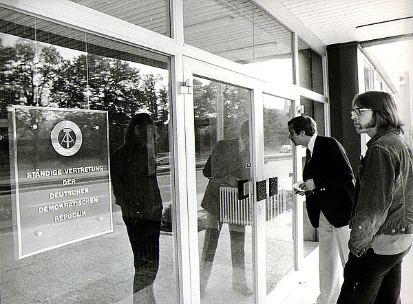 Zwei Männer stehen vor einer gläsernen Tür und schauen interessiert in das Gebäude hinein. An der Tür ist das Schild der Ständigen Vertretung der DDR in der Bundesrepublik Deutschland angebracht.
