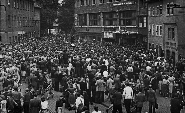 Schwarz-Weiß-Foto einer großen Menschenmenge auf einem Platz, der von Gebäuden umgeben ist.