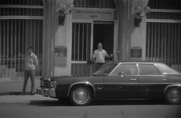 Schwarzweißfoto, zu sehen ist eine Person die aus einem Gebäude auf die Straße tritt. Vor ihr steht ein dunkles Auto. Rechts im Bild, an der Fassade des Gebäudes, steht eine Person die Uniform trägt.