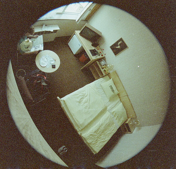 Mit Weitwinkelobjektv aufgenommenes Farbfoto eines Raums. Das Foto wurde aus der oberen Ecke des Zimmers gemacht. Oben rechts im Bild ist eine auf einem Stuhl sitzende Person zu sehen, die Zeitung liest. Oben mittig ist ein Fernseher, in der Bildmitte ist ein Bett zu sehen.