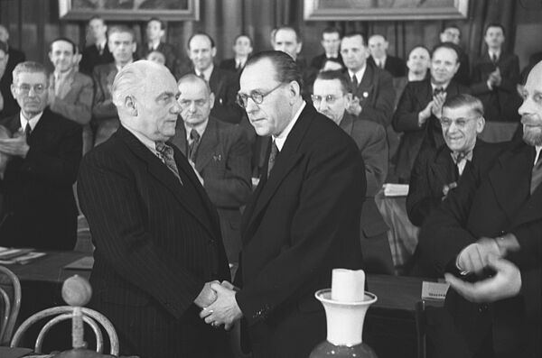 Das Schwarz-Weiß-Foto zeigt im Vordergrund zwei Männer im Anzug, die gegenseitig die Hände umklammern. Im Hintergrund sind mehrere Männer im Anzug, die applaudieren.