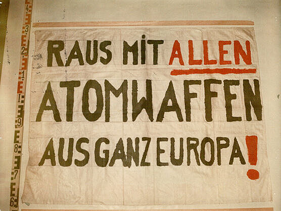 Beweisfoto der Stasi eines Transparents aus weißem Stoff. links am Bildrand ein Metermaß. Auf dem Transparent eine Aufschrift in schwarzen und teilweise roten Druckbuchstaben: "Raus mit allen Atomwaffen aus ganz Europa!"