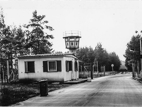 Blick die Zufahrtsstraße zum Stasi-Gelände in Freienbrink hinunter: links der Straße ein hüttenartiges Wärterhäuschen, dahinter ein Wachturm.