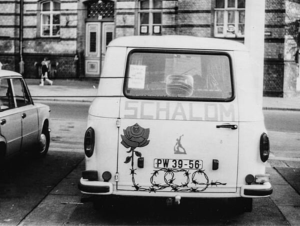 Foto eines weißen Kleinbusses auf dem "Schalom" steht, der Aufkleber einer Rose, deren Stiel aus Stacheldraht ist, sowie das "Schwerter zu Pflugscharen"-Symbol klebt