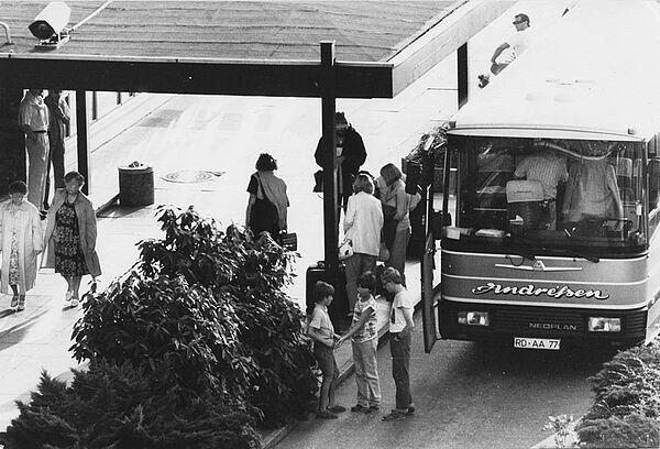 Schwarz-Weiß-Foto, das einen überdachten Eingangsbereich zu einem Gebäude zeigt. Davor befinden sich diverse Personengruppen, im vorderen Bildteil stehen drei Kinder neben einem Reisebus. Daneben stehen Menschen mit Koffern.