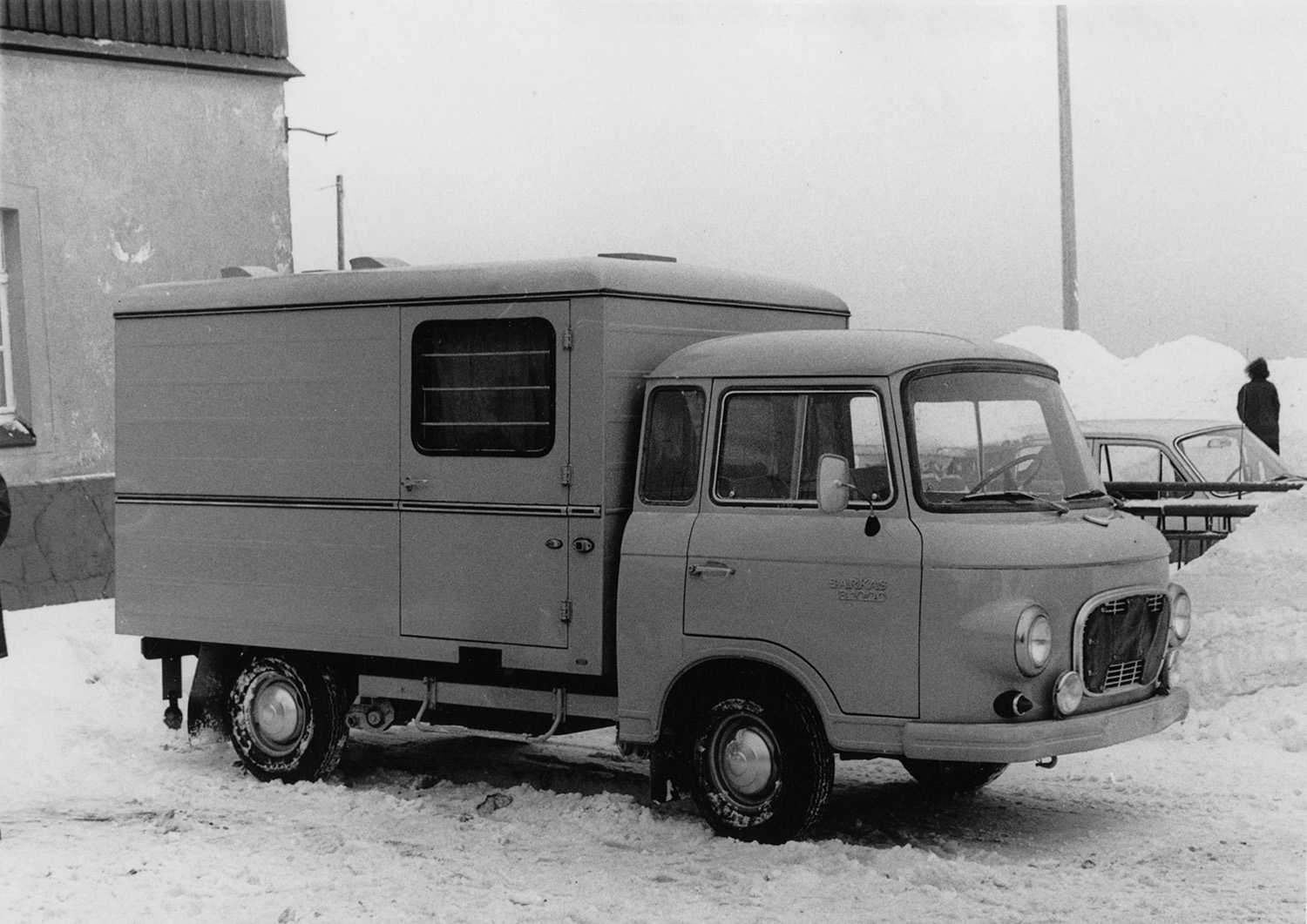 Gefangenentransportwagen (GTW), Typ Barkas 1000, o. D.; BArch, MfS, BV Karl-Marx-Stadt, AKG, Fo, Nr. 264, Bild 7 Schwarz-Weiß-Foto eines Kastenwagens bzw. Transporter, der vor einem Gebäude auf einem verschneiten Parkplatz steht.