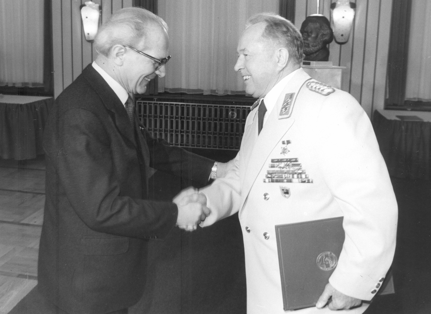 Honecker und Mielke schüttel sich freudig lächelnd die Hände. Honecker trägt einen schwarzen Anzug, Mielke eine weiße Galauniform.