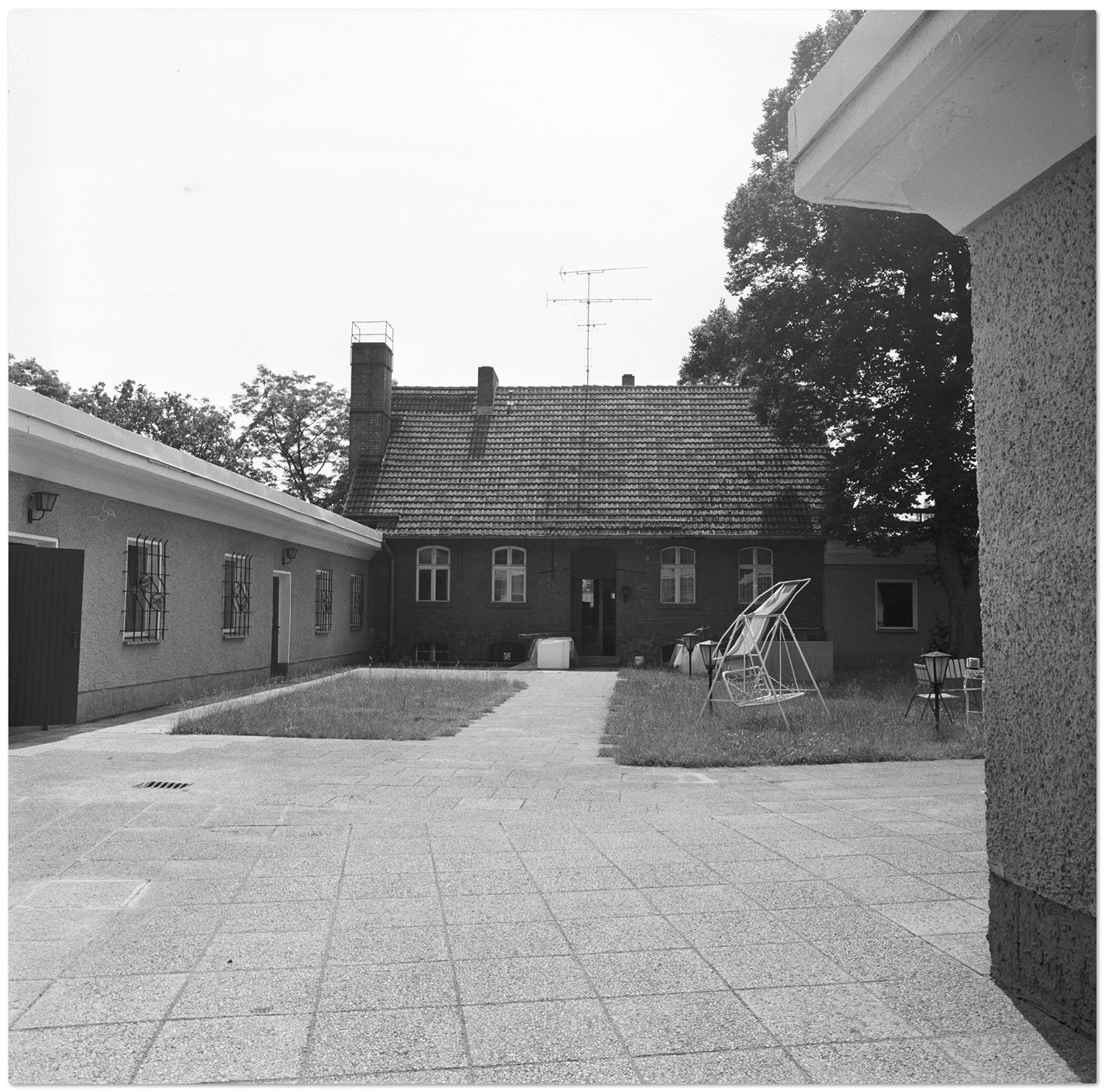Innenhof des konspirativen „Objekts 74“ In Briesen, wo die Stasi internationale Terroristen unterbrachte5 Blick in einen Hof. Im Hintergrund ein aus roten Ziegeln errichtetes Wohnhaus. Im Vordergrund eine mit Platten gepflasterte Fläche. Dazwischen eine Rasenfläche auf der eine Hollywood-Schaukel steht. links davon ein eingeschossiger Neubau-Gebäudetrakt. Rechts im Vordergrund ist ein Teil der Außenwand eines weiteren Gebäudes zu sehen.