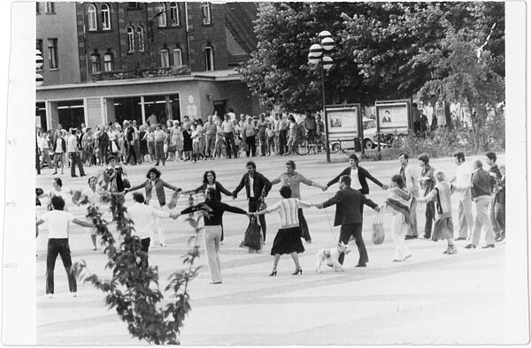 Schwarz-Weiß-Foto:Eine Gruppe von Menschen tanzt händehaltend auf einem Platz im Kreis. Im Hintergrund hat sich eine Menschenmenge zum Zuschauen gebildet.