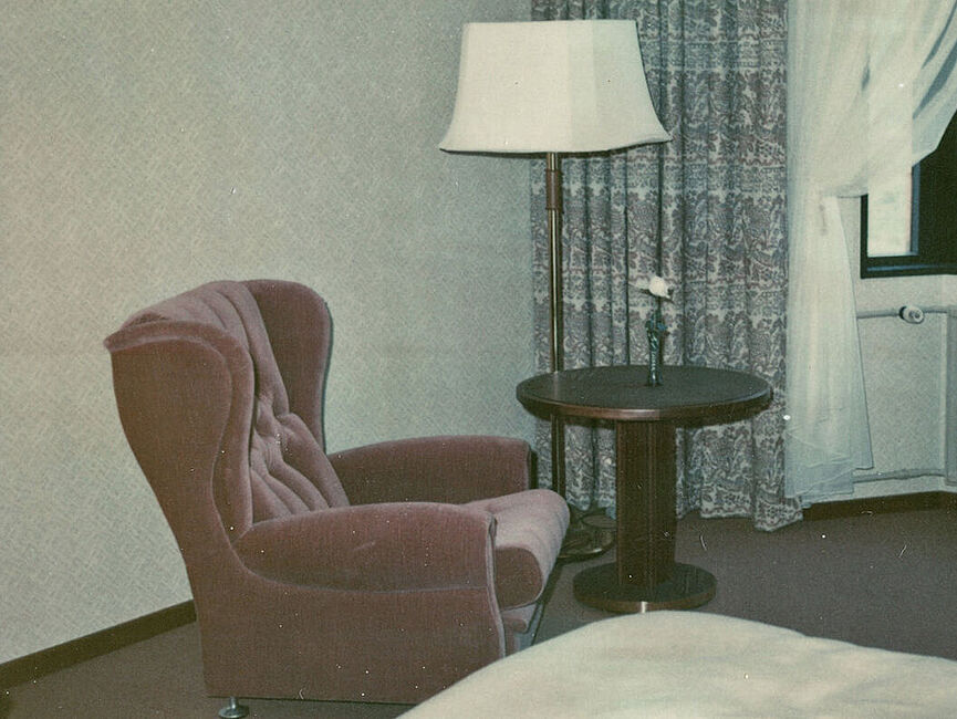 Aufnahmen der Einrichtungsgegenstände eines Musterzimmers im Interhotel "Bellevue" aus einer Dokumentation der Stasi zur Fernsprechanlage mit Leitungsnetz im Hotelneubau Farbfoto eines Hotelzimmers, im Vordergrund ist ein Bett zu sehen, im Hintergrund ein Sessel und ein Stuhl, dazwischen ein Couchtisch, dahinter eine Lampe und ein Fenster.