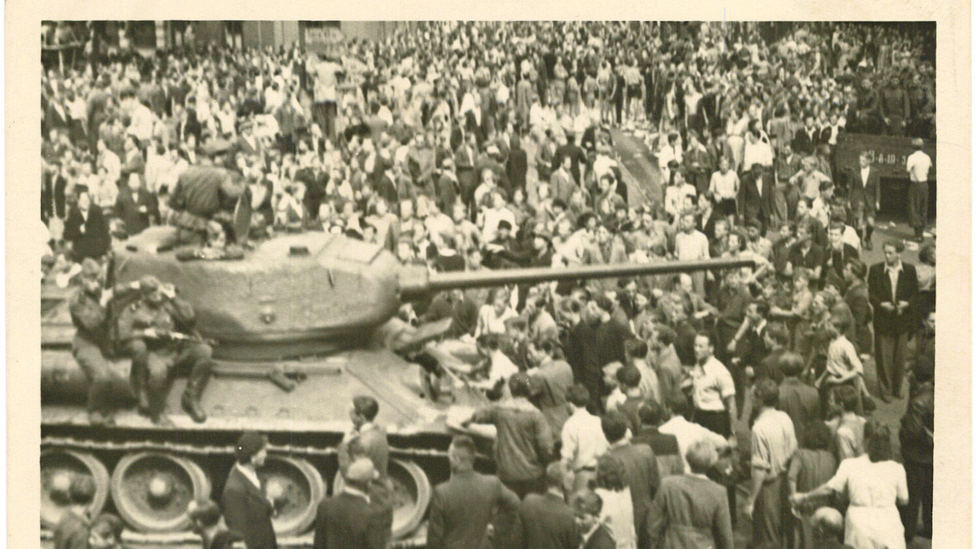 Schwarz-Weiß-Foto einer Menschenmenge, die einen Panzer umringt, auf dem bewaffnete Soldaten sitzen.