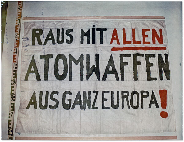 Beweisfoto der Stasi eines Transparents aus weißem Stoff. links am Bildrand ein Metermaß. Auf dem Transparent eine Aufschrift in schwarzen und teilweise roten Druckbuchstaben: "Raus mit allen Atomwaffen aus ganz Europa!"