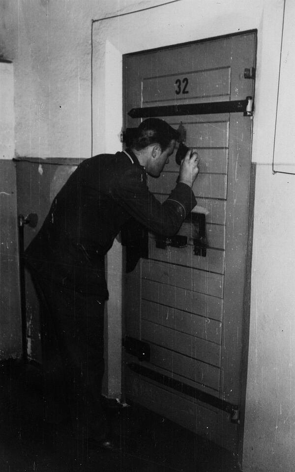 Foto aus einer Stasi-Dokumentation über Einrichtung und Sicherungsmaßnahmen in Haftanstalten, Quelle: BArch, MfS, Abt. XIV, Fo, Nr. 69, Bl. 6, Bild 7 Schwarz-Weiß-Foto eines unifmorieten Mannes, der durch einen Schlüsselspion in eine Zellentür schaut.
